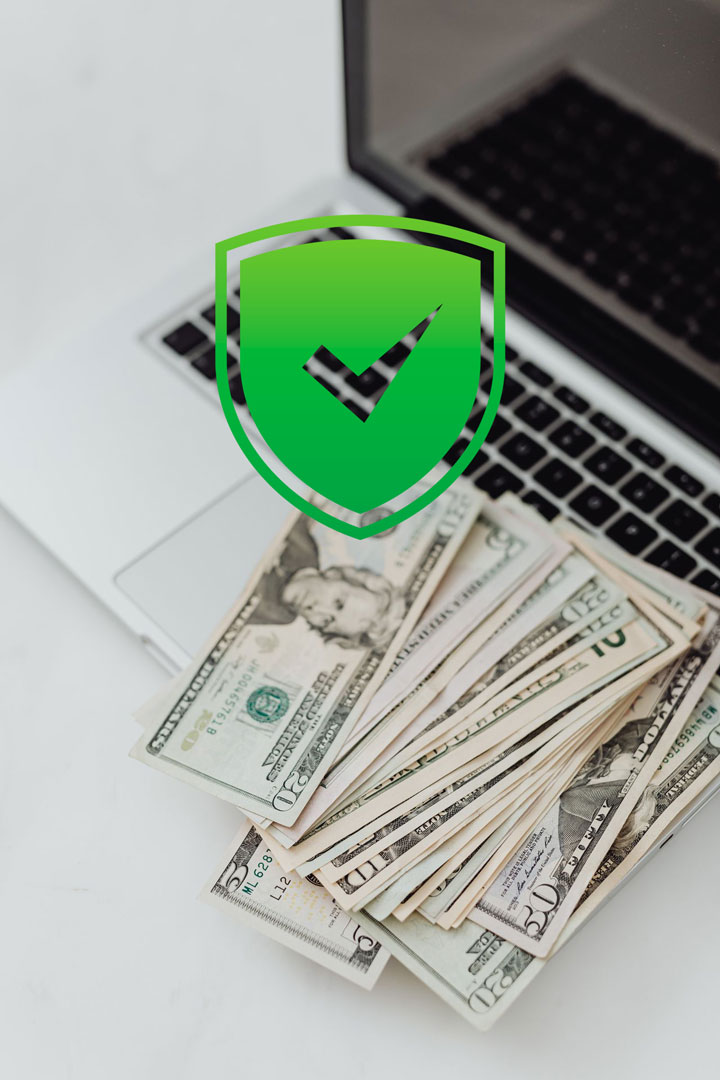 A stack of US dollar bills is spread out on a laptop keyboard, with a large green security shield and checkmark icon digitally superimposed over the image.