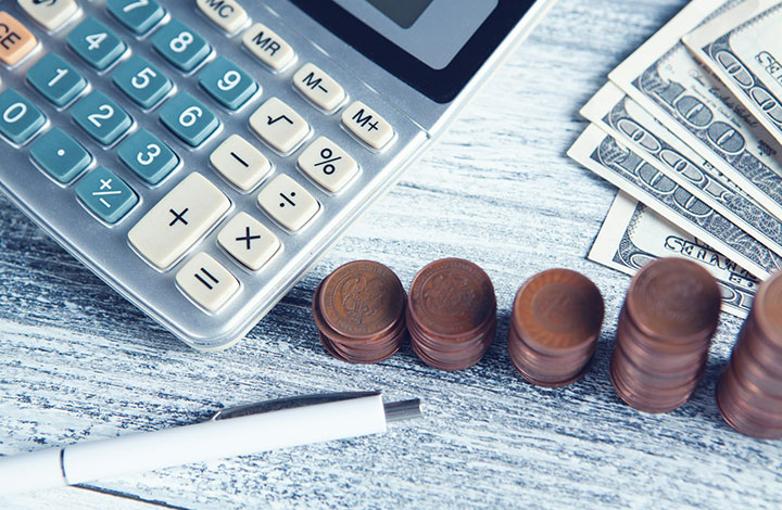 A close-up of a calculator, stacks of coins, a white pen, and several US dollar bills on a light wooden surface, representing financial planning or budgeting.