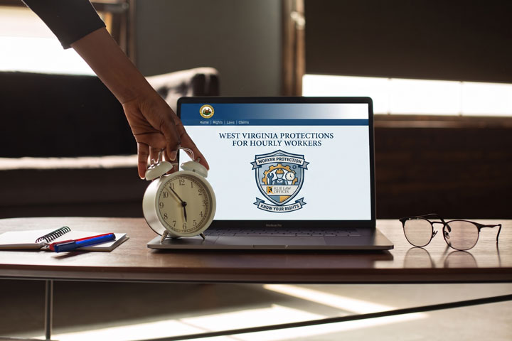 A hand sets a white alarm clock on a table in front of a laptop displaying "West Virginia Protections for Hourly Workers." On the table are a notepad with a pen and a pair of eyeglasses.