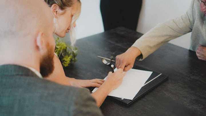 Three people sit around a dark table, with one person signing a document while another points at the paper, and a third watches. A small plant is partially visible in the background.