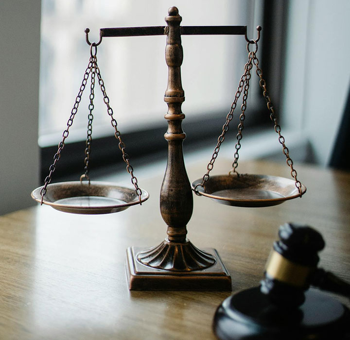A bronze balance scale sits on a wooden desk beside a judge’s gavel, with a window and urban buildings blurred in the background.