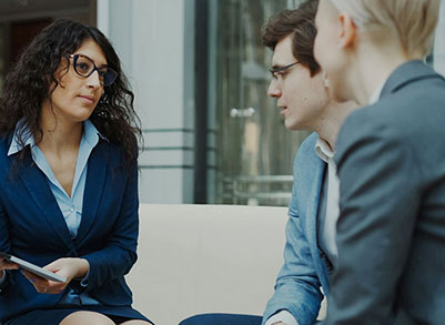 Three people in business attire sit together indoors, engaged in a serious discussion. One woman with dark curly hair and glasses holds a tablet, while the other two, a man and a woman, listen attentively.