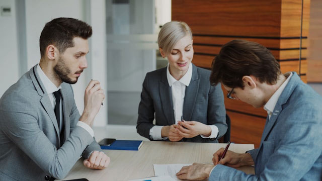 Three people in business attire sit at a table in an office. One person is signing a document while the other two, a man and a woman, watch and engage in conversation.