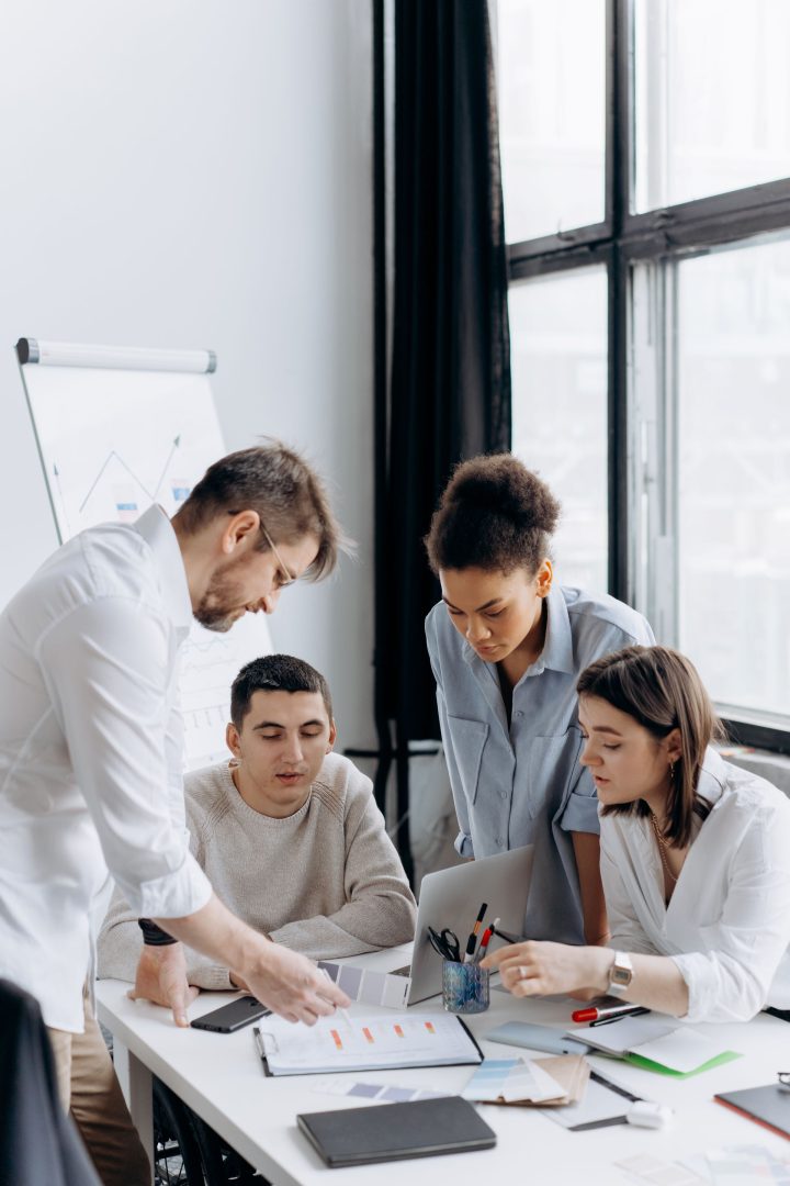 Four people are gathered around a desk in a bright office, discussing documents and charts. One person is pointing at a paper while others look on attentively, with a laptop and office supplies on the table.