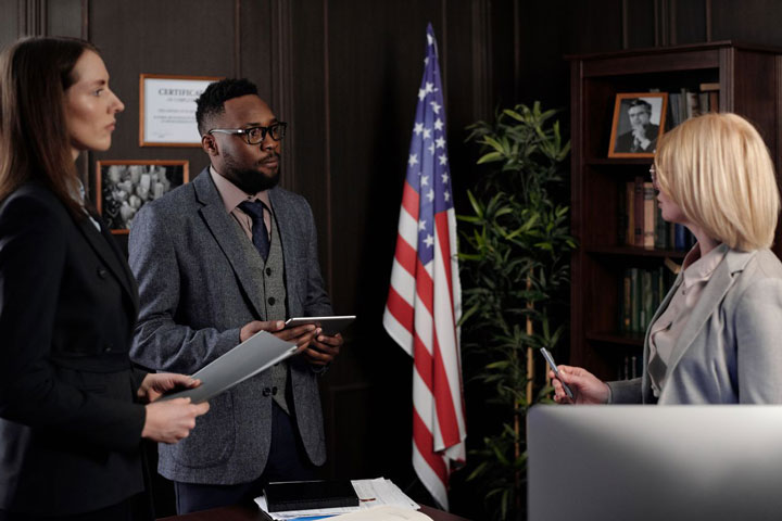 Three professionals in business attire have a discussion in an office with an American flag, shelves with books, and framed photos in the background. Two hold tablets while one speaks, holding a pen.