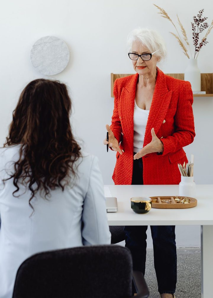 An older woman in a red blazer gestures while speaking to another woman seated at a desk in a modern office, with decor and office supplies visible.