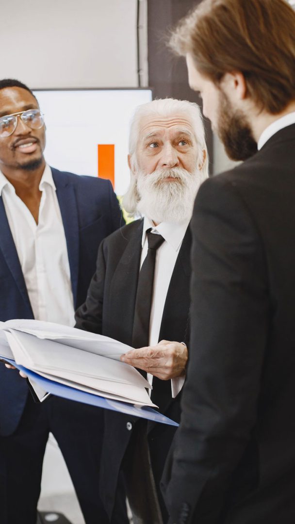 Three men in suits are having a discussion, with one older man holding and showing documents to the others. They appear to be in a professional or business setting.
