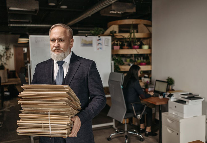 An older man in a suit holds a large stack of files in an office, while a woman works at a desk with a computer in the background.