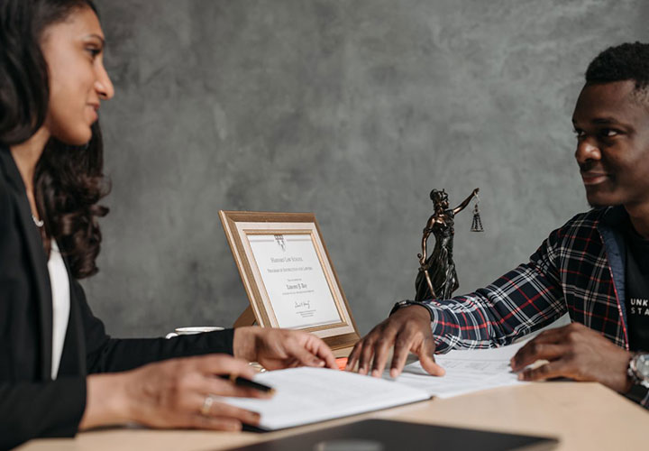 Two people sit at a desk reviewing documents. A framed certificate and a small Lady Justice statue are on the desk. They appear to be in a professional, possibly legal, setting.