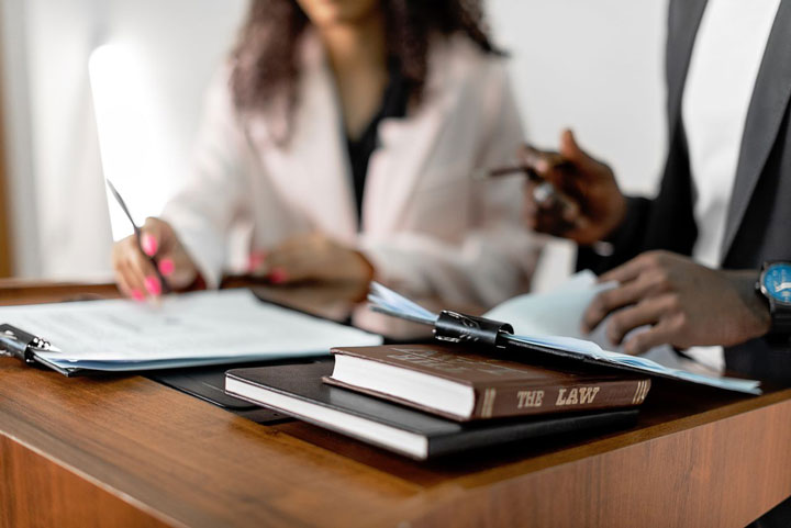 Two people in business attire work at a desk with documents, pens, and a book titled “The Law” in focus in the foreground.