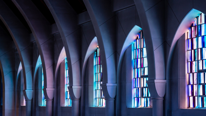 Tall arched windows with colorful stained glass panels line a church wall, casting blue and purple light inside a vaulted, modern interior.