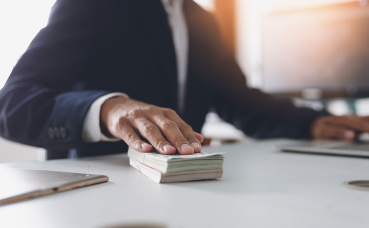 A person in a suit sits at a desk with one hand resting on a stack of cash, using a laptop with the other hand.