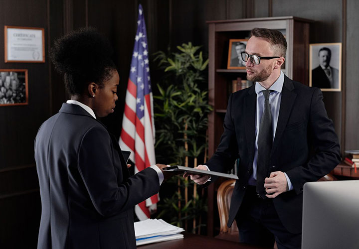 Two people in business suits talk in an office. One hands a file to the other. An American flag, framed photos, and office plants are visible in the background.