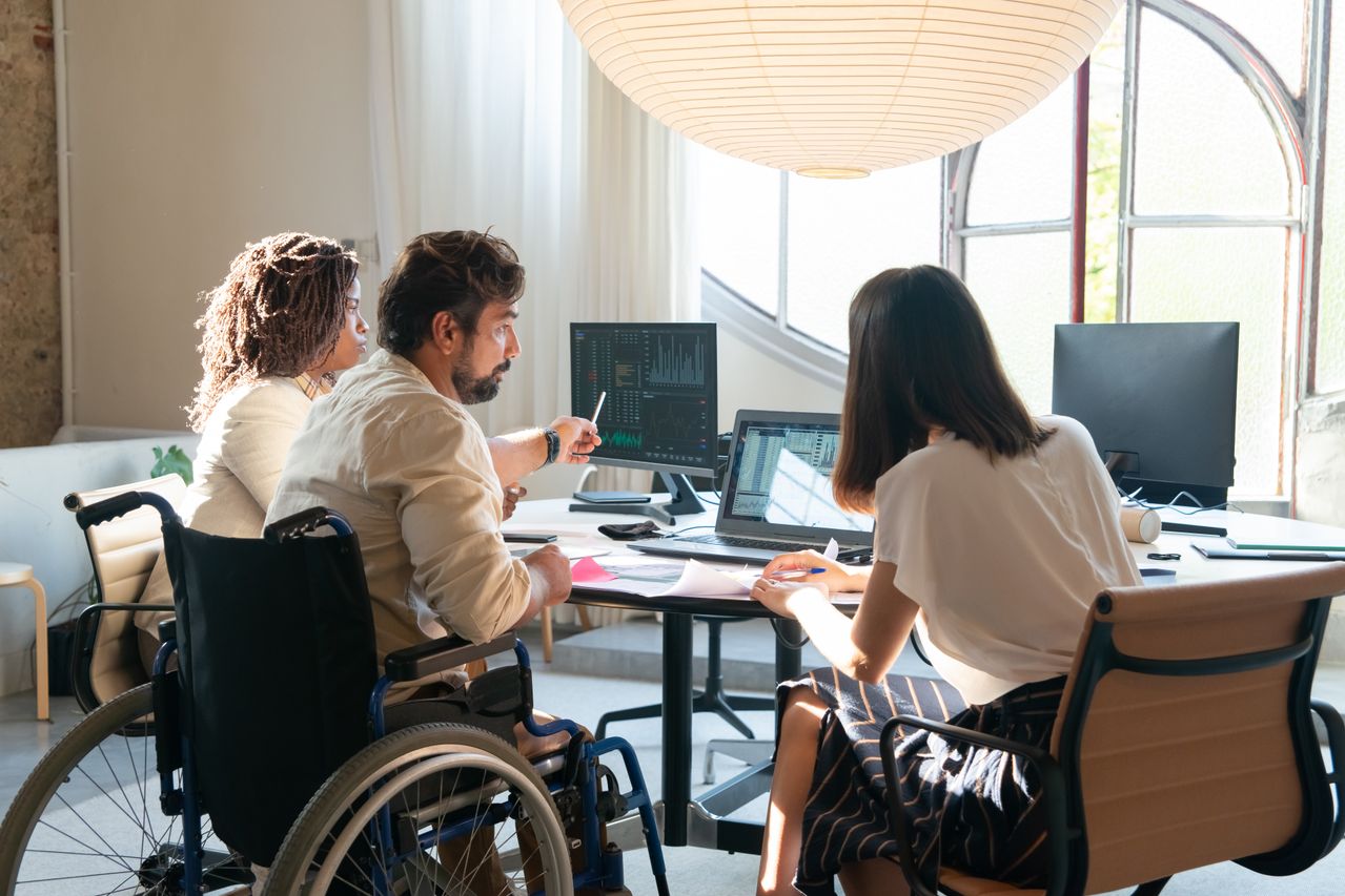 Three people, one in a wheelchair, discuss data at a table with laptops and monitors in a bright office. Papers are spread out, and one person points at the screen showing charts.
