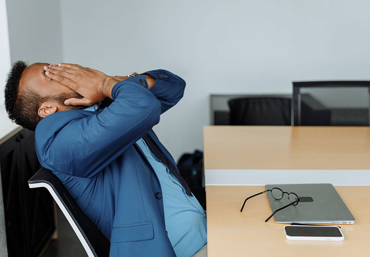 A person in a blue suit sits at a desk, leaning back with hands covering their face. Glasses, a laptop, and a phone are on the table, suggesting stress or frustration in an office setting.
