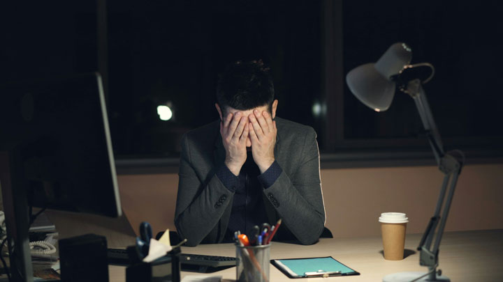 A person in a suit sits at a desk at night, covering their face with their hands, surrounded by a computer, lamp, coffee cup, and office supplies, suggesting stress or exhaustion.