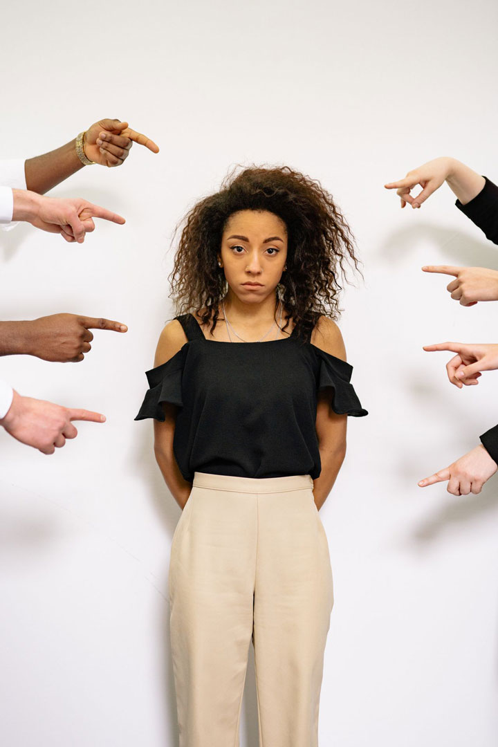 A woman stands against a white wall, looking serious, while multiple hands from different directions point at her, suggesting blame or accusation.