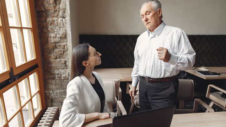 An older man in a white shirt talks to a seated woman in a blazer by a window in an office, while she looks up at him. A laptop sits on the table in front of her.