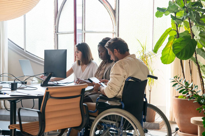 Three people are working together at a desk in a bright office. One person is in a wheelchair, and all are focused on their laptops and notes. There are large windows and indoor plants in the background.