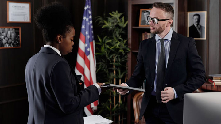 A woman hands a folder to a man in an office with bookshelves and an American flag in the background. Both wear business suits and appear to be in a serious discussion.