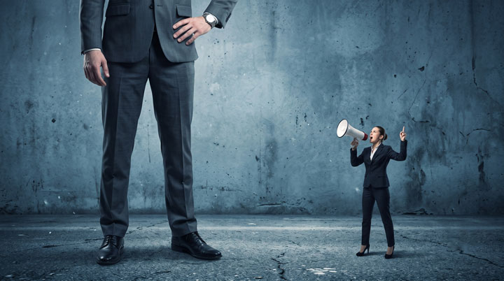A giant man in a suit stands confidently while a much smaller woman in business attire shouts through a megaphone at him against a gray, textured wall.