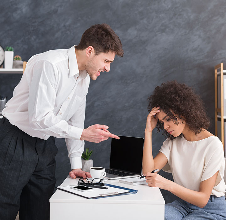 A man in a white shirt angrily points at a woman sitting at a desk with her head in her hand, looking stressed. Papers, a laptop, and a cup are on the desk.