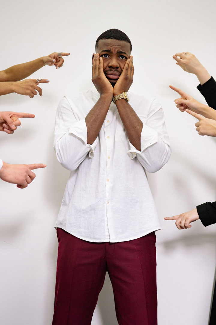 A man stands with a worried expression, holding his face, while multiple hands around him point accusingly.