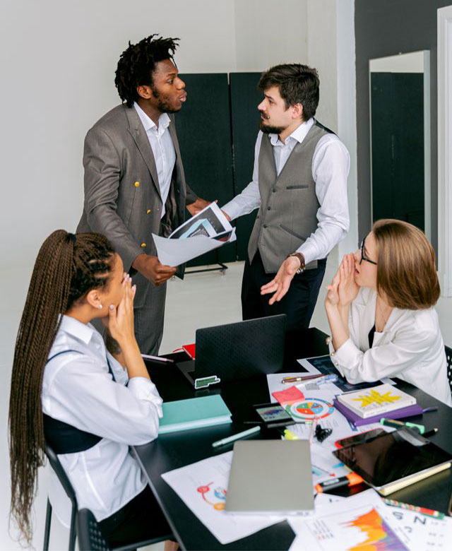 Two men in suits have a serious discussion while two women sit at a desk with charts and laptops, looking engaged. The workspace is modern, with papers, notebooks, and a cup of coffee on the table.