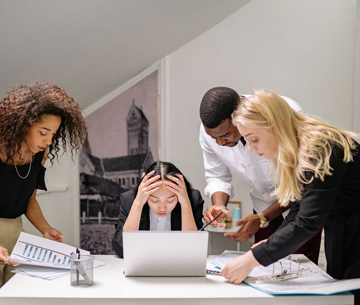 Four coworkers gather around a desk with papers and a laptop. One person sits with their head in their hands, looking stressed, while the others stand, discussing and pointing at documents.