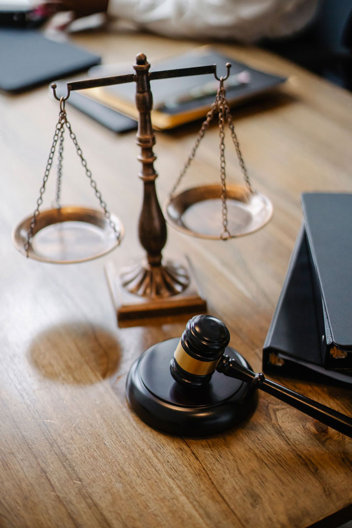 A judge’s gavel and a set of brass balance scales rest on a wooden table, with black folders nearby, suggesting a legal or courtroom setting.