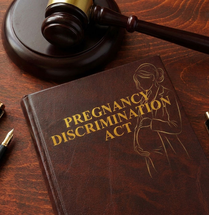 A book titled "Pregnancy Discrimination Act" with an outline of a pregnant woman on the cover, next to a judge's gavel and a pen on a wooden table.
