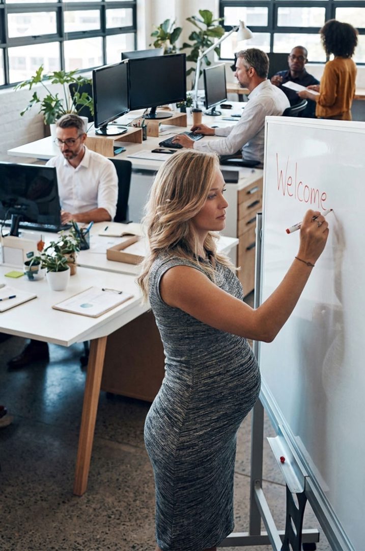 A pregnant woman writes "Welcome" on a whiteboard in a modern office with several people working at desks and computers in the background.