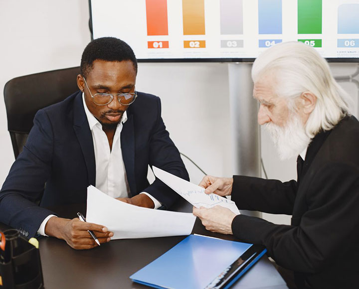 Two men in business suits review documents at a desk, with charts and graphs displayed on a screen in the background. One man is younger with glasses, the other is older with white hair and beard.