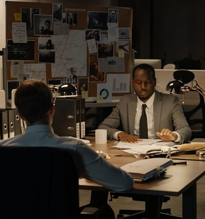 Two men sit at a desk covered in papers and folders, discussing documents. Behind them is a bulletin board with maps, photos, and notes, suggesting an investigation or detective work atmosphere.