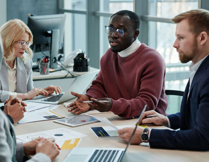 Four professionals sit around a table in an office, discussing documents and laptops. One man gestures while speaking, and others listen attentively, suggesting a business meeting or team collaboration.
