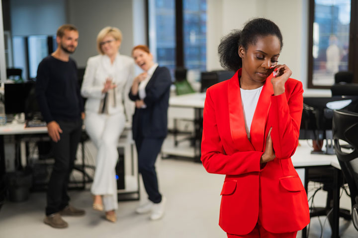 A woman in a red suit looks upset in an office, while three colleagues in the background stand together, appearing to gossip and laugh.