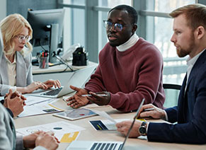 Four people sit at a table with laptops and papers, engaged in a discussion in a modern office setting. One person is speaking while others listen and take notes.