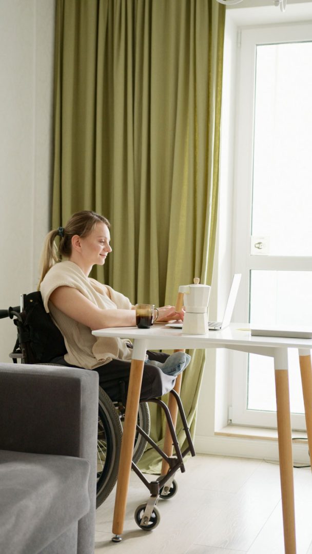 A woman in a wheelchair works on a laptop at a table near a window with green curtains. There is a white mug and teapot on the table.