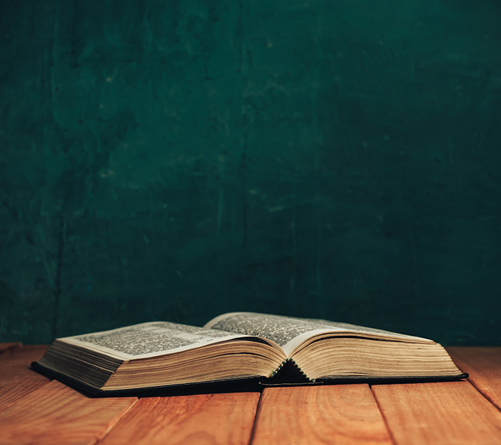 An open book rests on a wooden table with a dark green textured wall in the background. The book pages are fanned out, and the scene appears warmly lit.