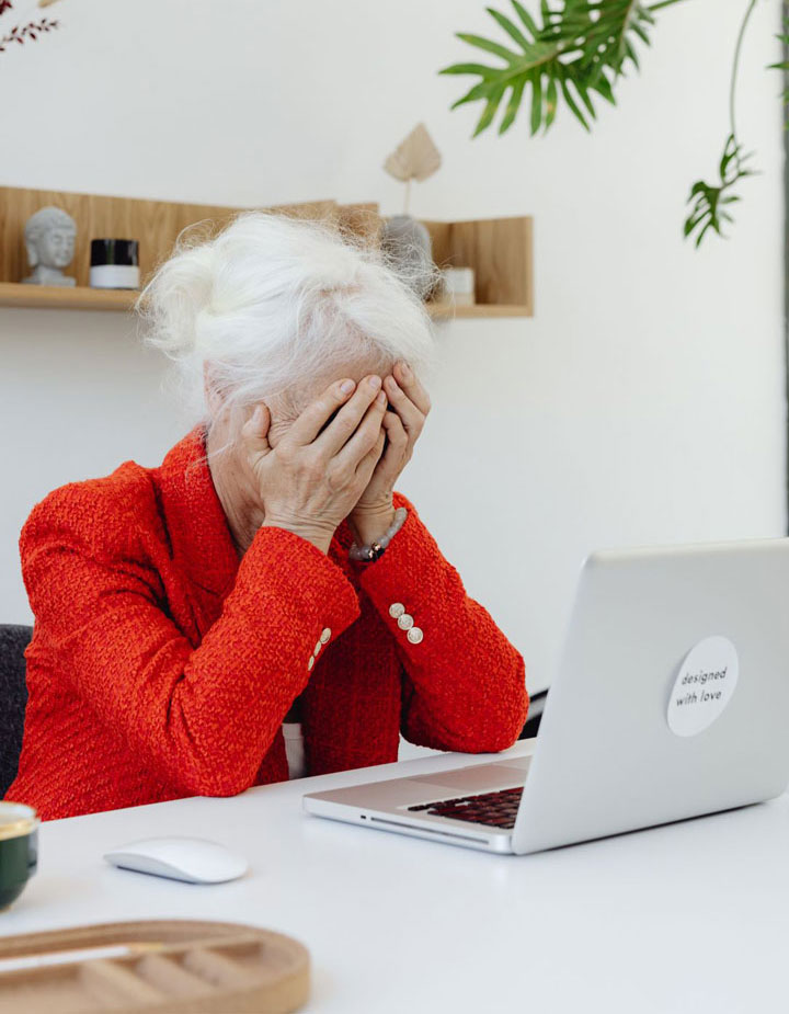 An elderly woman in a red jacket sits at a desk with her head in her hands, looking stressed in front of a laptop.