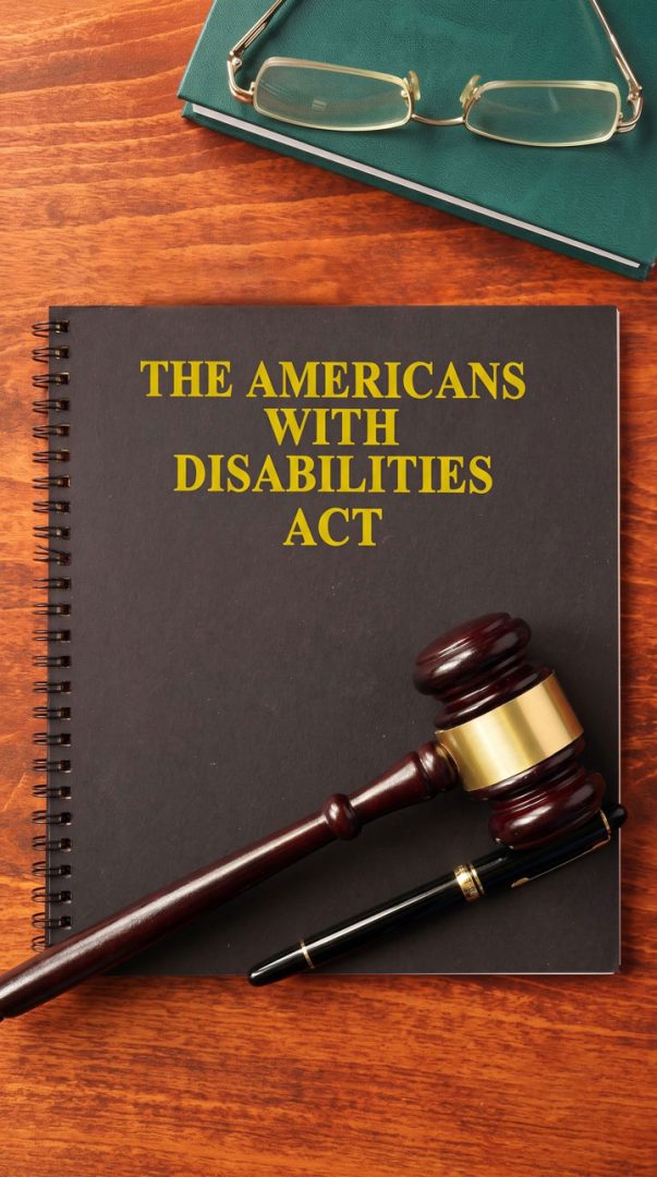 A notebook titled "The Americans with Disabilities Act" sits on a wooden desk with a gavel, pen, and a closed folder with glasses on top.
