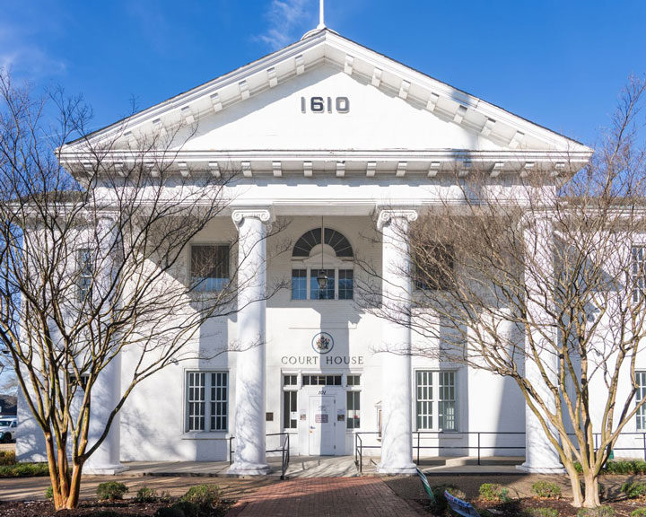 White courthouse with four tall columns, a triangular pediment displaying "1610," and leafless trees in front. The building has large windows and a central door, all under a bright blue sky.