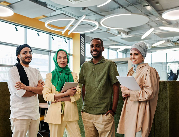Four young professionals stand together in a modern office, smiling at the camera. Two women wear headscarves and hold tablets, while two men stand beside them, one with arms crossed. Contemporary decor is visible in the background.