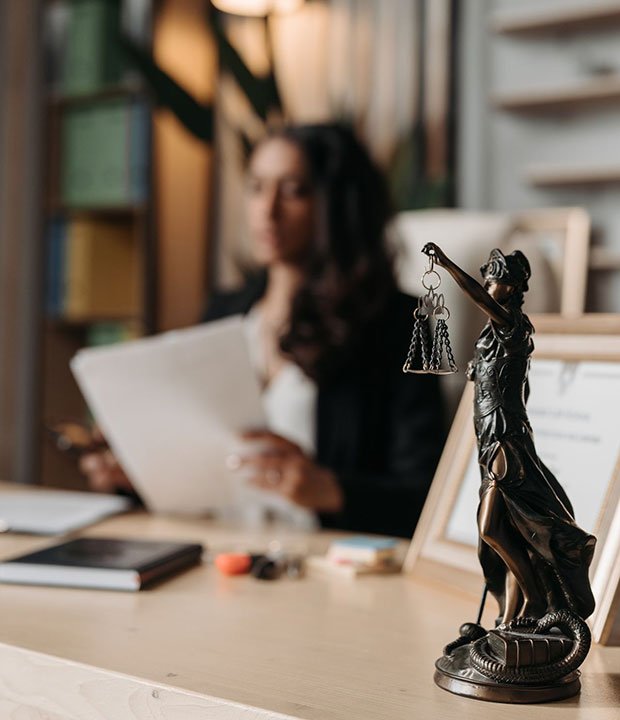 A statuette of Lady Justice stands on a desk in focus, while a woman holding papers sits blurred in the background in an office setting.