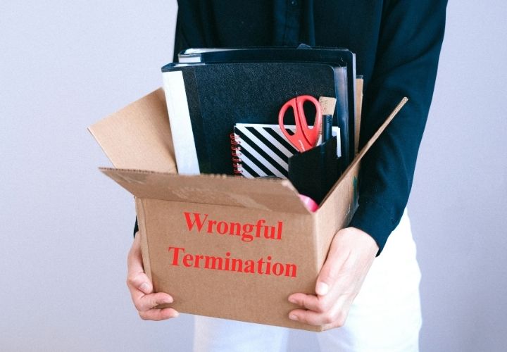 A person in a black shirt holds a cardboard box filled with office supplies, including folders and red scissors. The box has "Wrongful Termination" printed on the front in red letters.