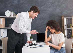 A man is standing and pointing angrily at a seated woman who looks stressed and is holding her head in one hand and paperwork in the other. They are in an office with shelves and a clock in the background.