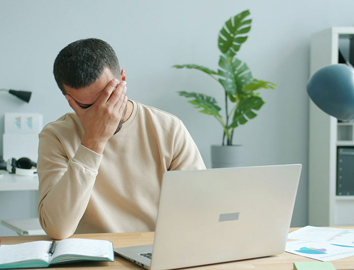 A person sits at a desk with a laptop, covering their face with one hand as if stressed or frustrated. There are papers and a notebook on the desk and a large plant in the background.