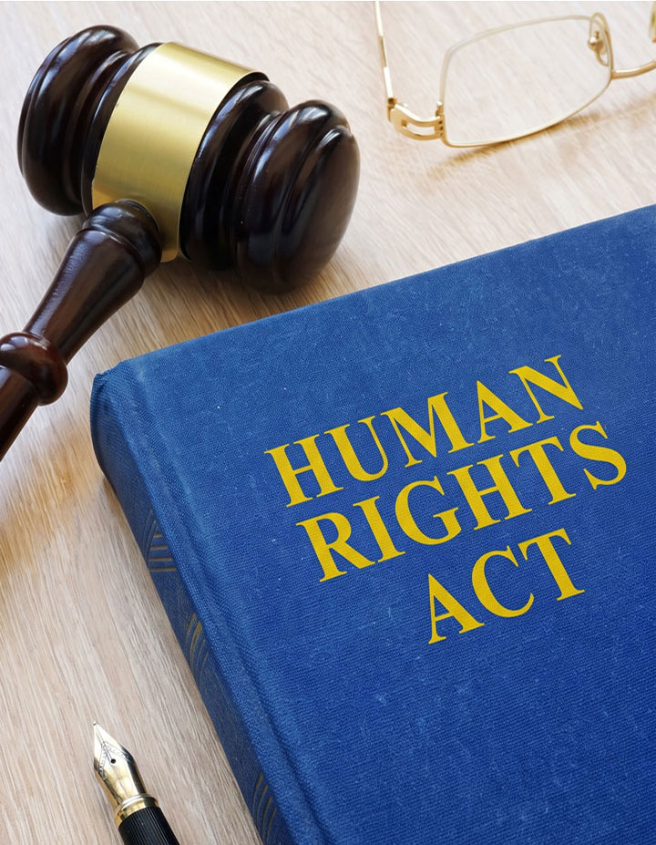 A blue book titled “HUMAN RIGHTS ACT” sits on a desk with a judge’s gavel, eyeglasses, and a pen nearby.