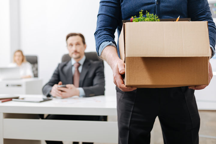 A person holding a cardboard box filled with office items stands in the foreground, while two people sit at desks in the background, one appearing serious.