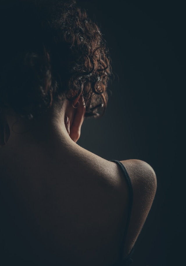 A person with curly hair is shown from behind, with their hand touching the side of their neck. The lighting is dim, highlighting the contours of their shoulder and neck against a dark background.